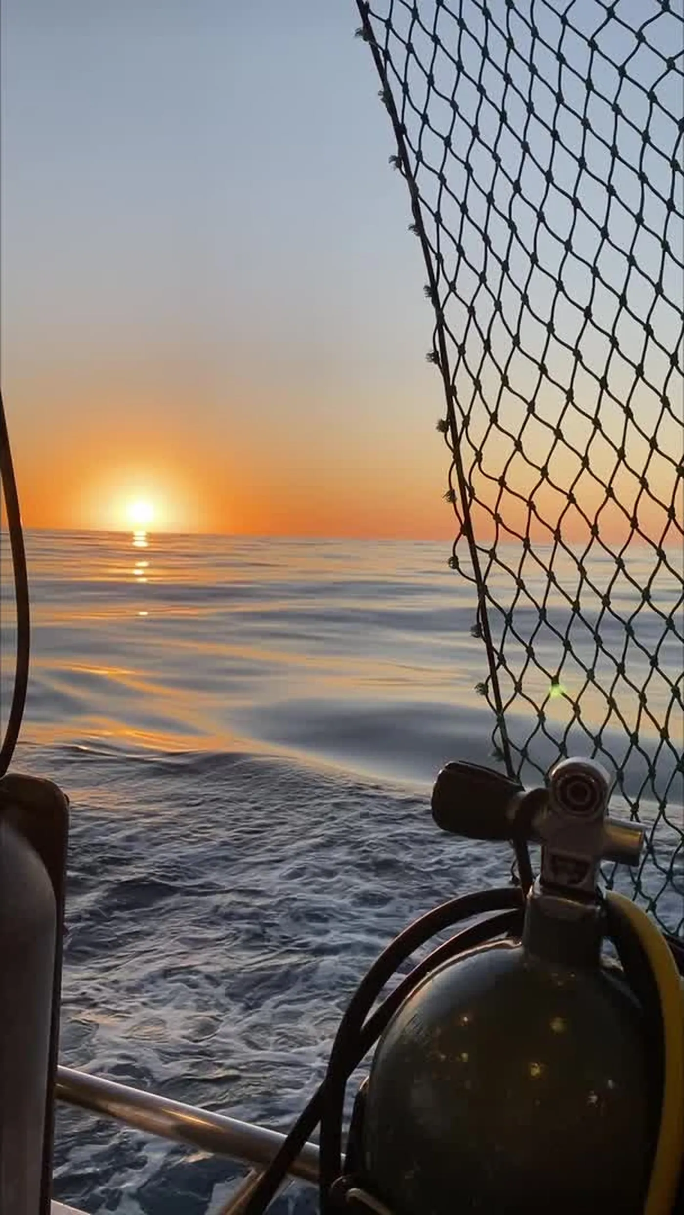 Sunrise over calm water seen from the deck of a dive boat, scuba tank in the foreground